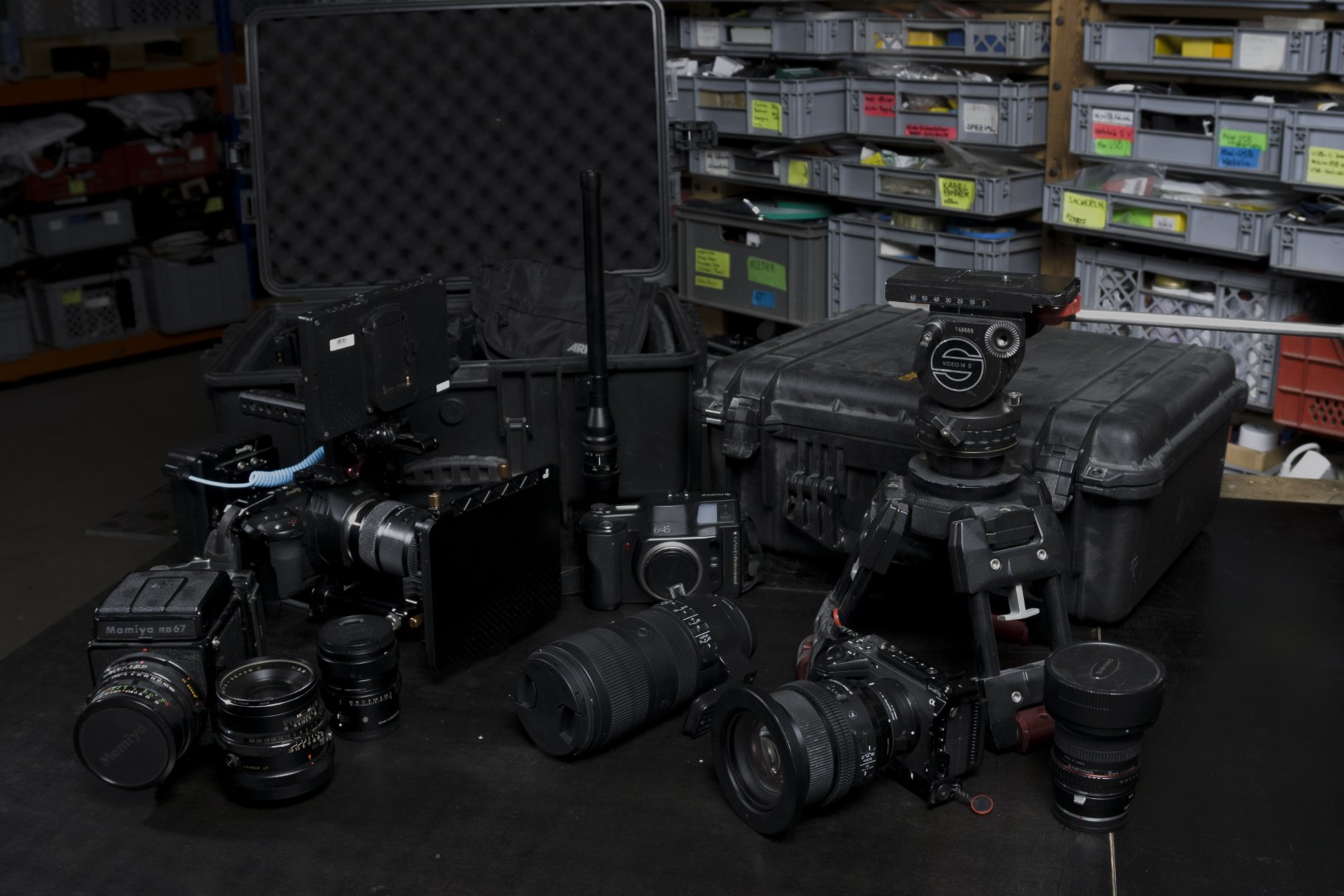 Professional camera equipment on a black table including multiple cameras, lenses, a camera tripod, and protective cases in a workshop setting.
