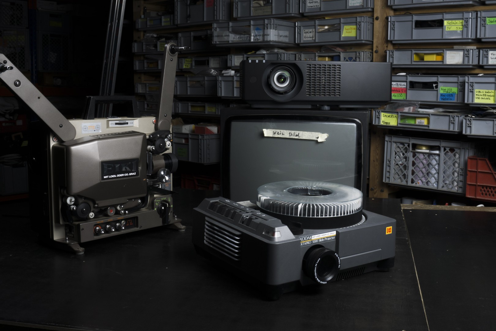 Vintage slide projectors and a small monitor on a table in front of shelves filled with labeled storage bins.
